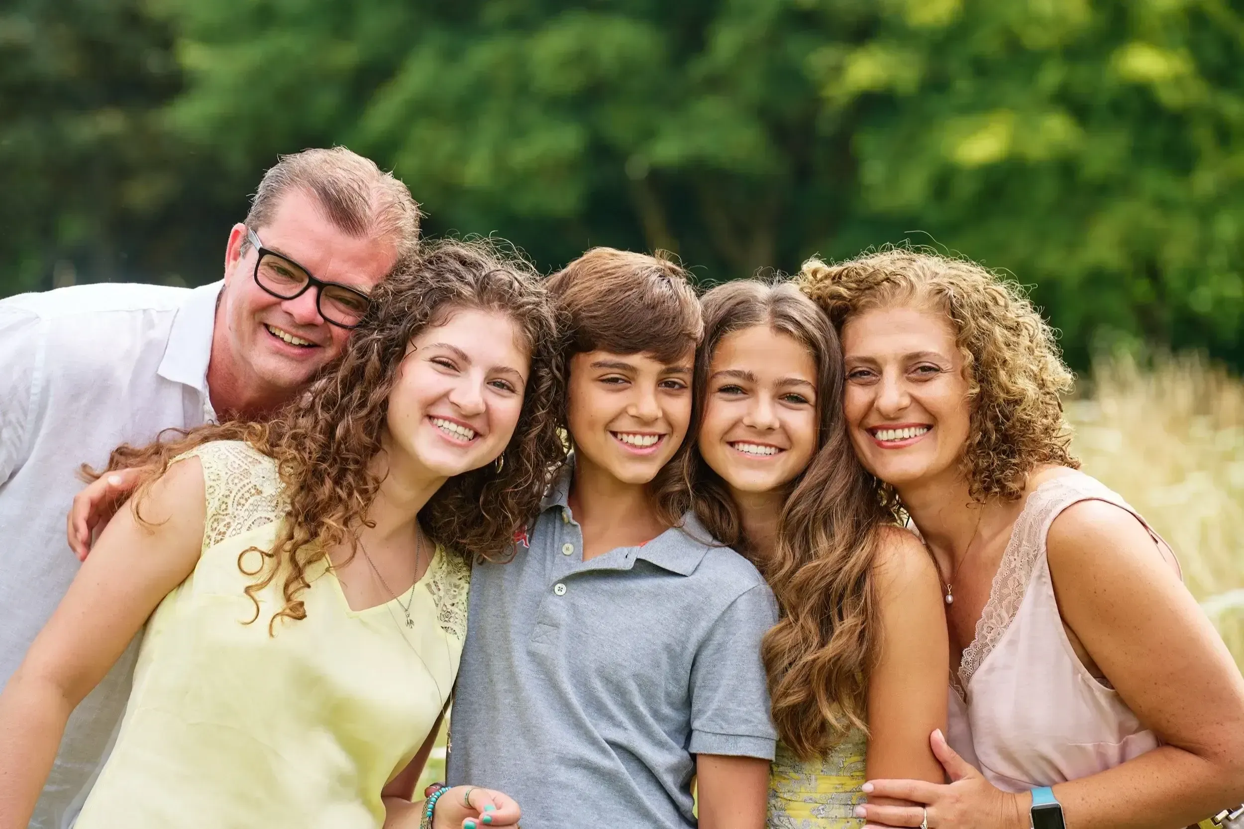 Een groep mensen, bestaande uit drie vrouwen en drie tienerkinderen, staat dicht bij elkaar en lacht buiten in een groene omgeving.