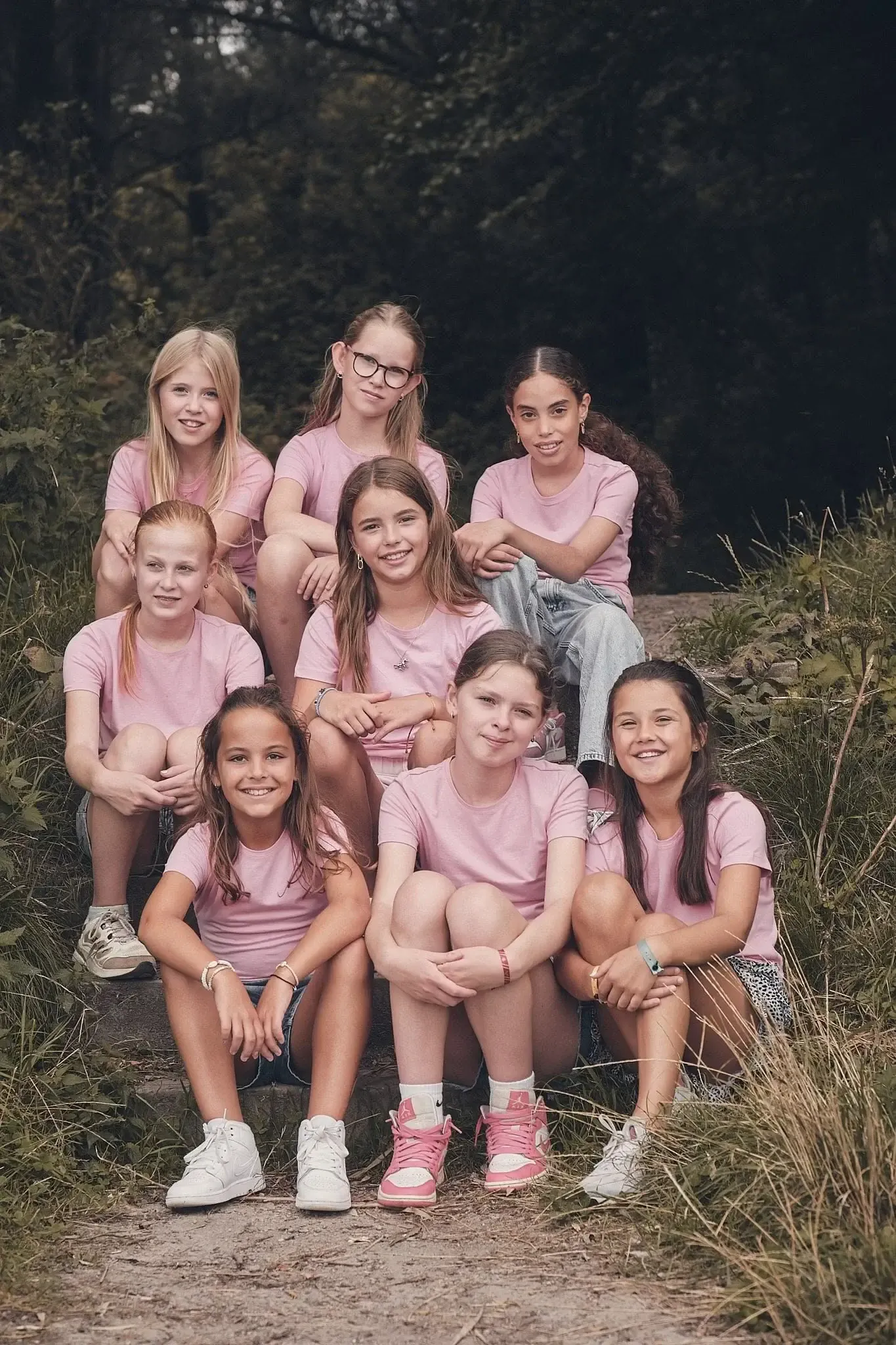 Tien meisjes in roze T-shirts en verschillende gekleurde sneakers zittend op een bosweggetje, omgeven door groen, lachen naar de camera.