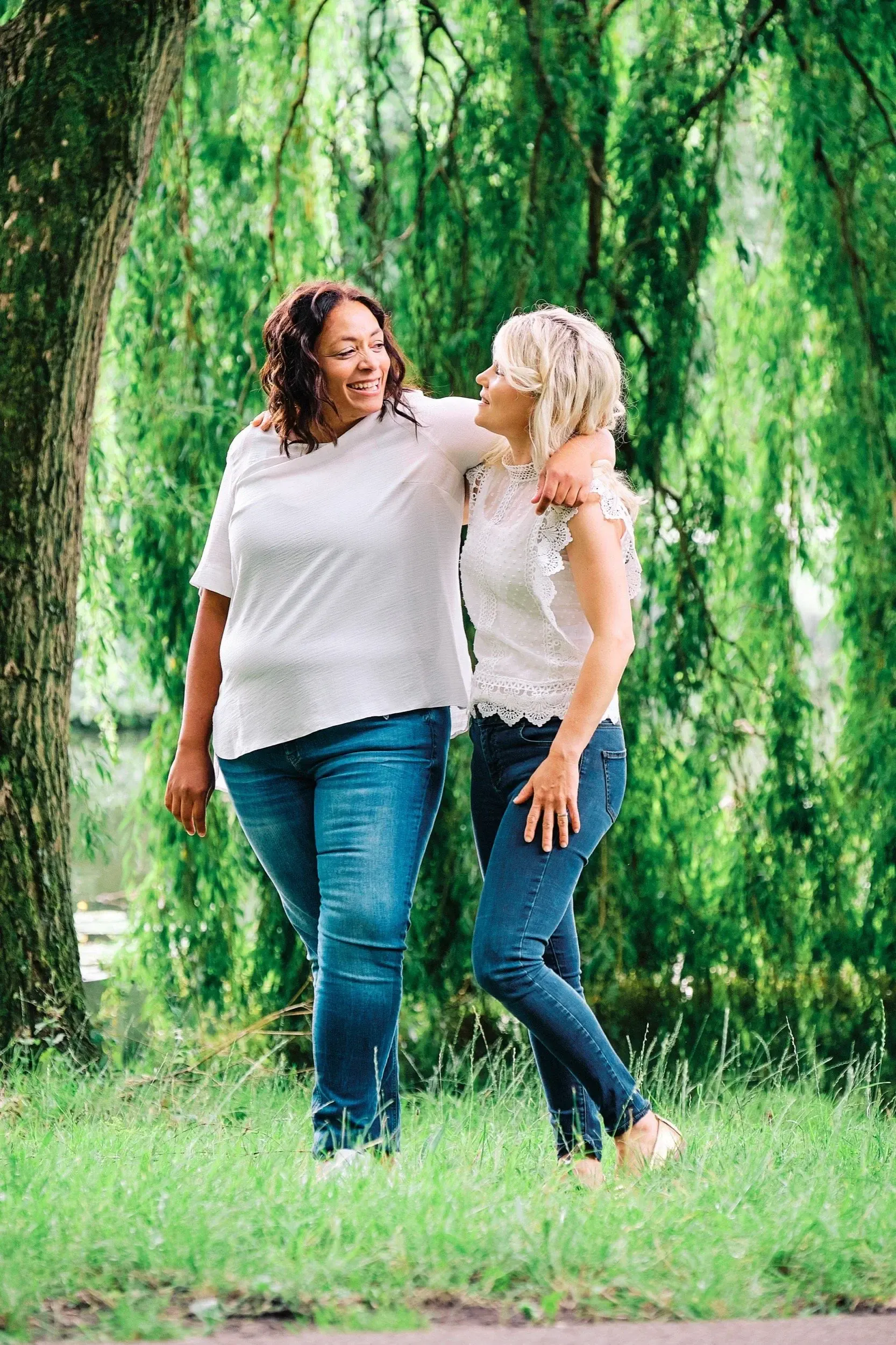 Twee vrouwen die samen door een groen bos lopen en vriendelijk naar elkaar glimlachen.
