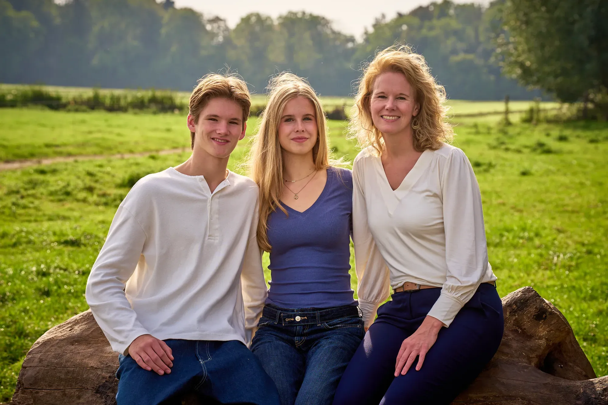 Moeder met tienerzoon en -dochter op een boomstam in een open veld, met zacht tegenlicht in de haren