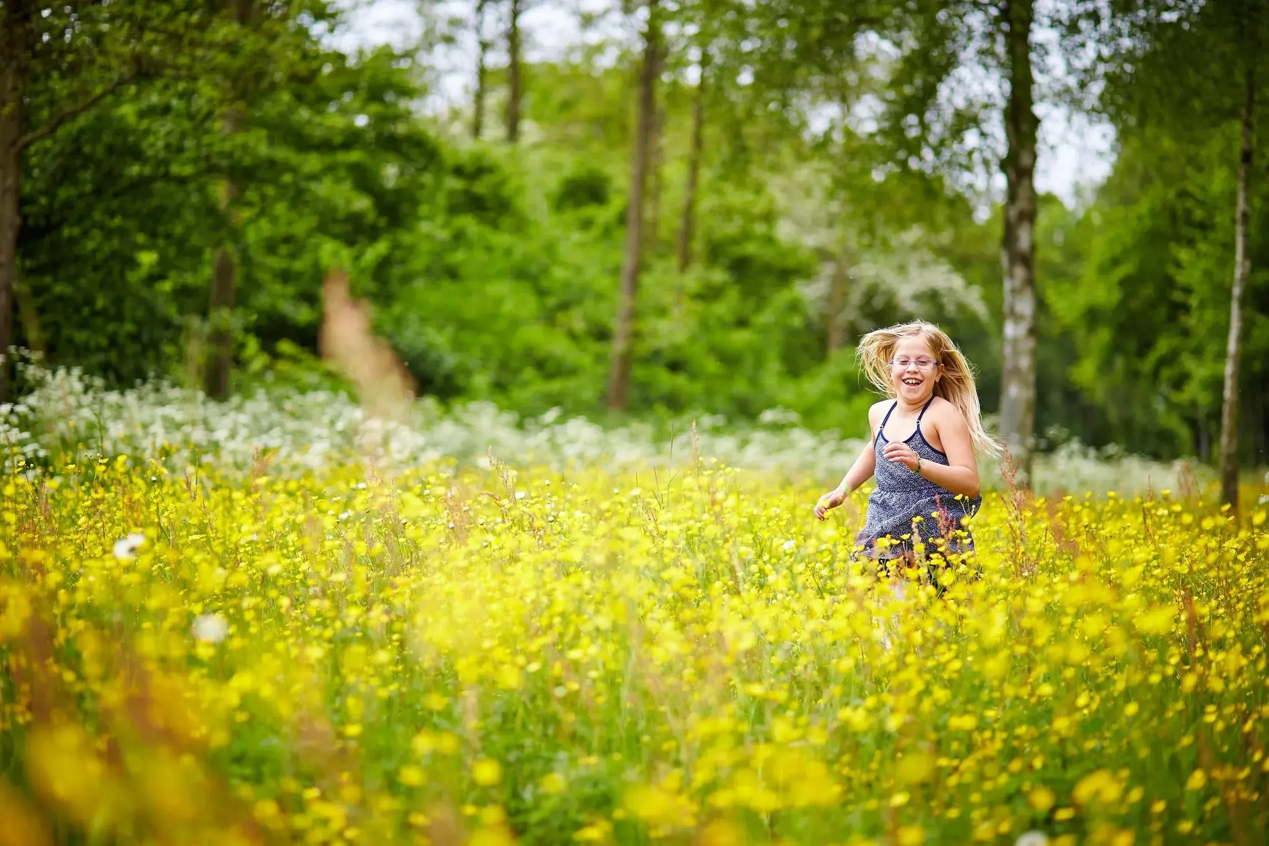 Een lachend jong meisje dat door een weide vol gele bloemen rent met een boswachterij op de achtergrond.