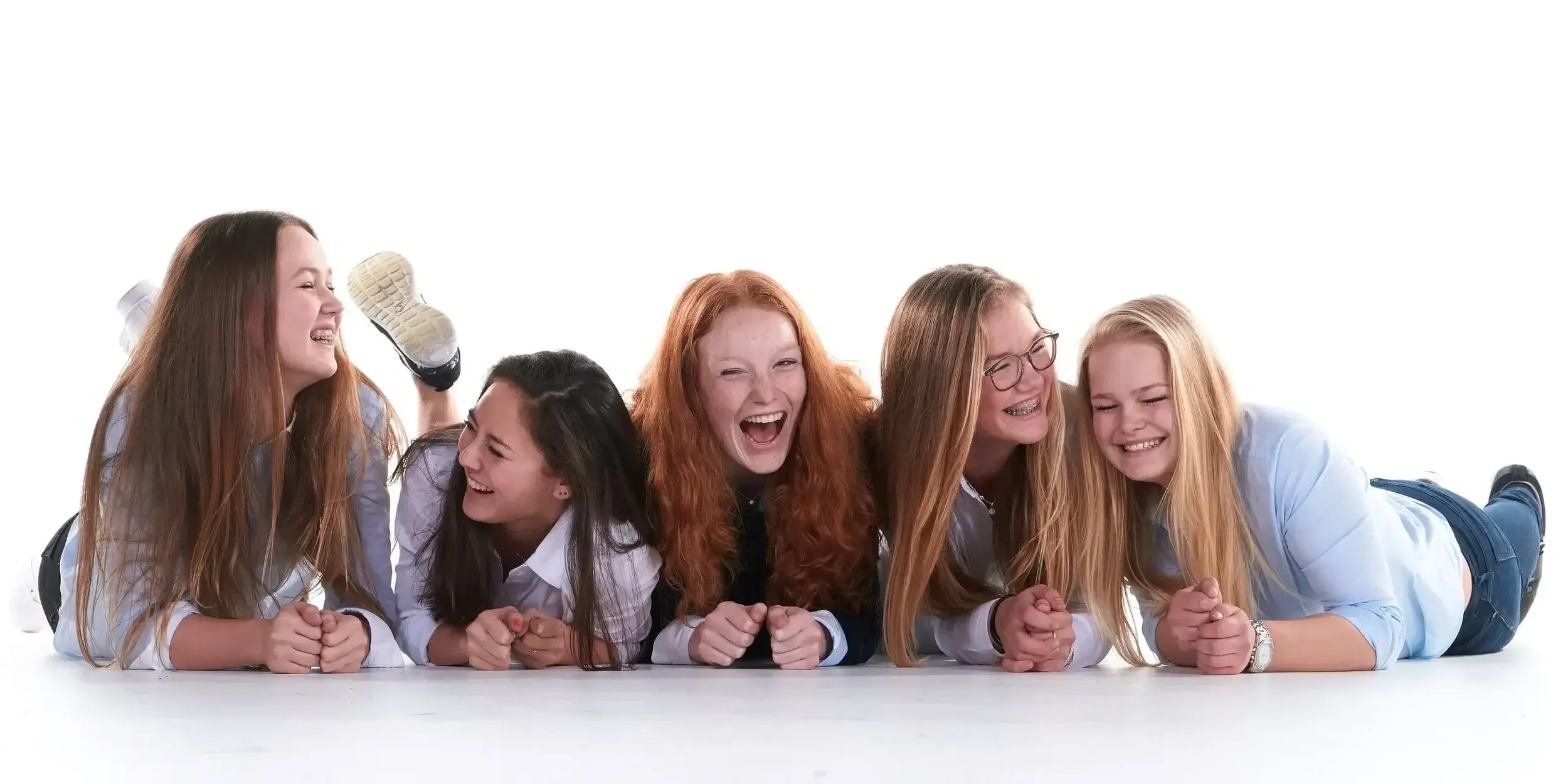 Vijf jonge vrouwen liggen op de buik op een witte achtergrond, lachen en praten samen.