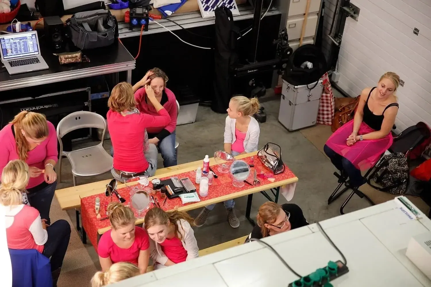 Verschillende vrouwen, onder wie een jonge meisje, zitten en staan in een ruimte met make-up en haaraccessoires, wellicht tijdens een modeshow of fotoshoot voorbereiding.