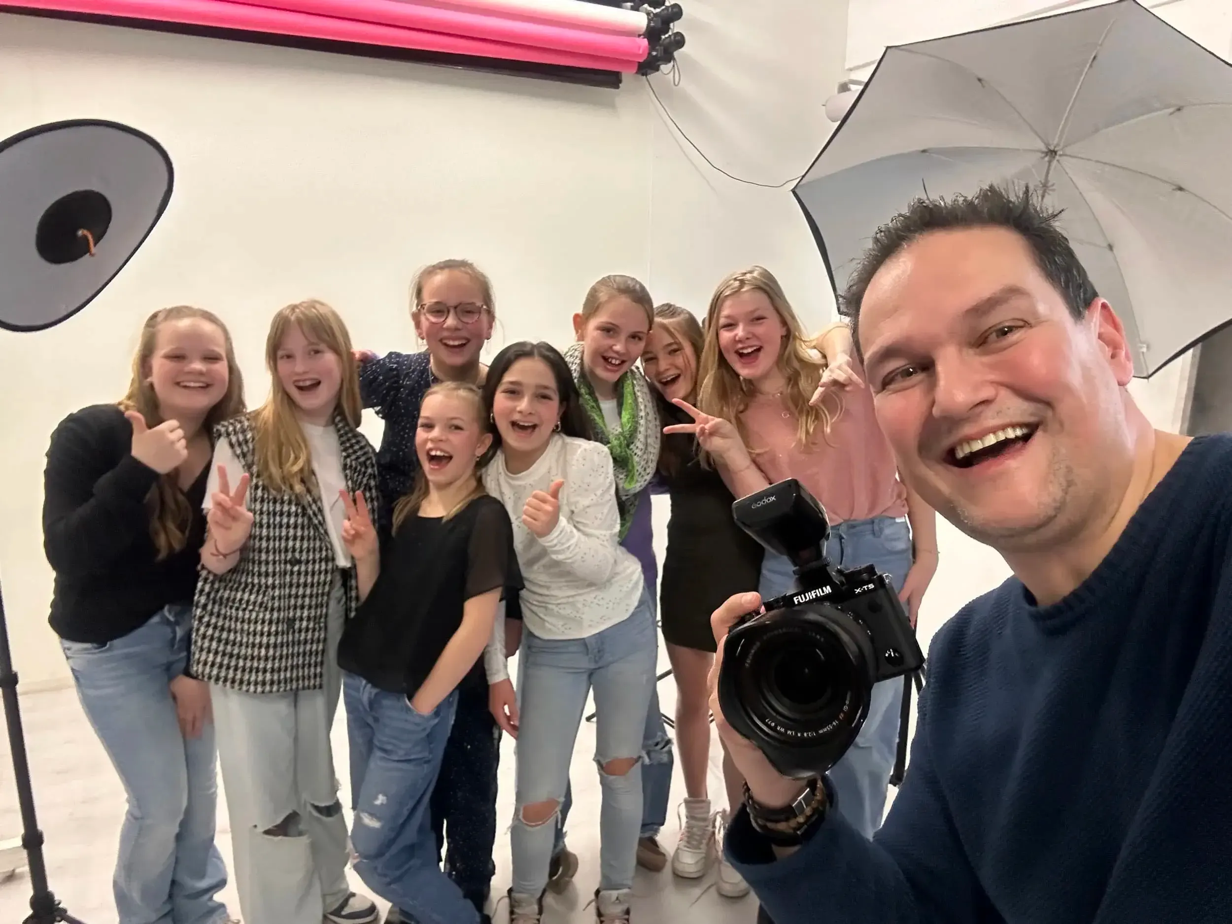 Een groep jonge meisjes poseert voor een foto in een studio, met een man die de camera vasthoudt. De meisjes lachen en maken V-vlaggen met hun handen.