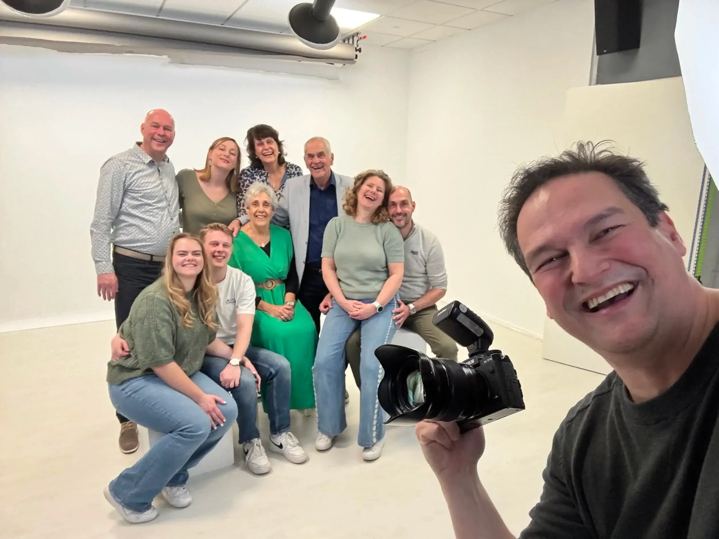 Groep mensen poseert voor een foto in een witte ruimte, de meeste mensen lachen en lijken gelukkig, een man houdt een camera vast en maakt een selfie.