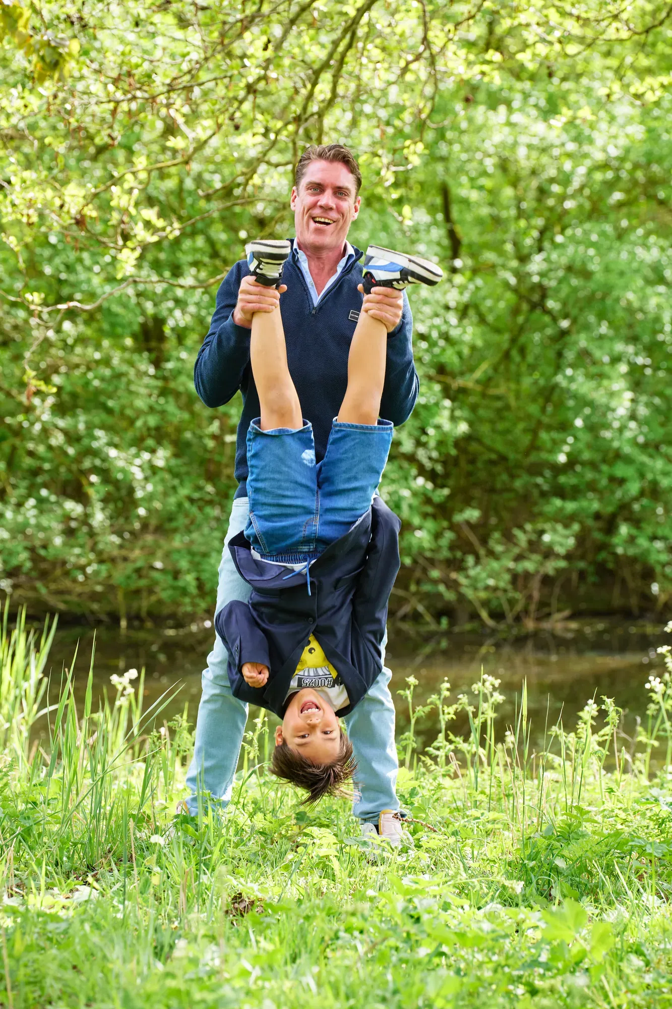 Vader houdt zijn zoon ondersteboven vast in het gras bij het water in het Westerpark