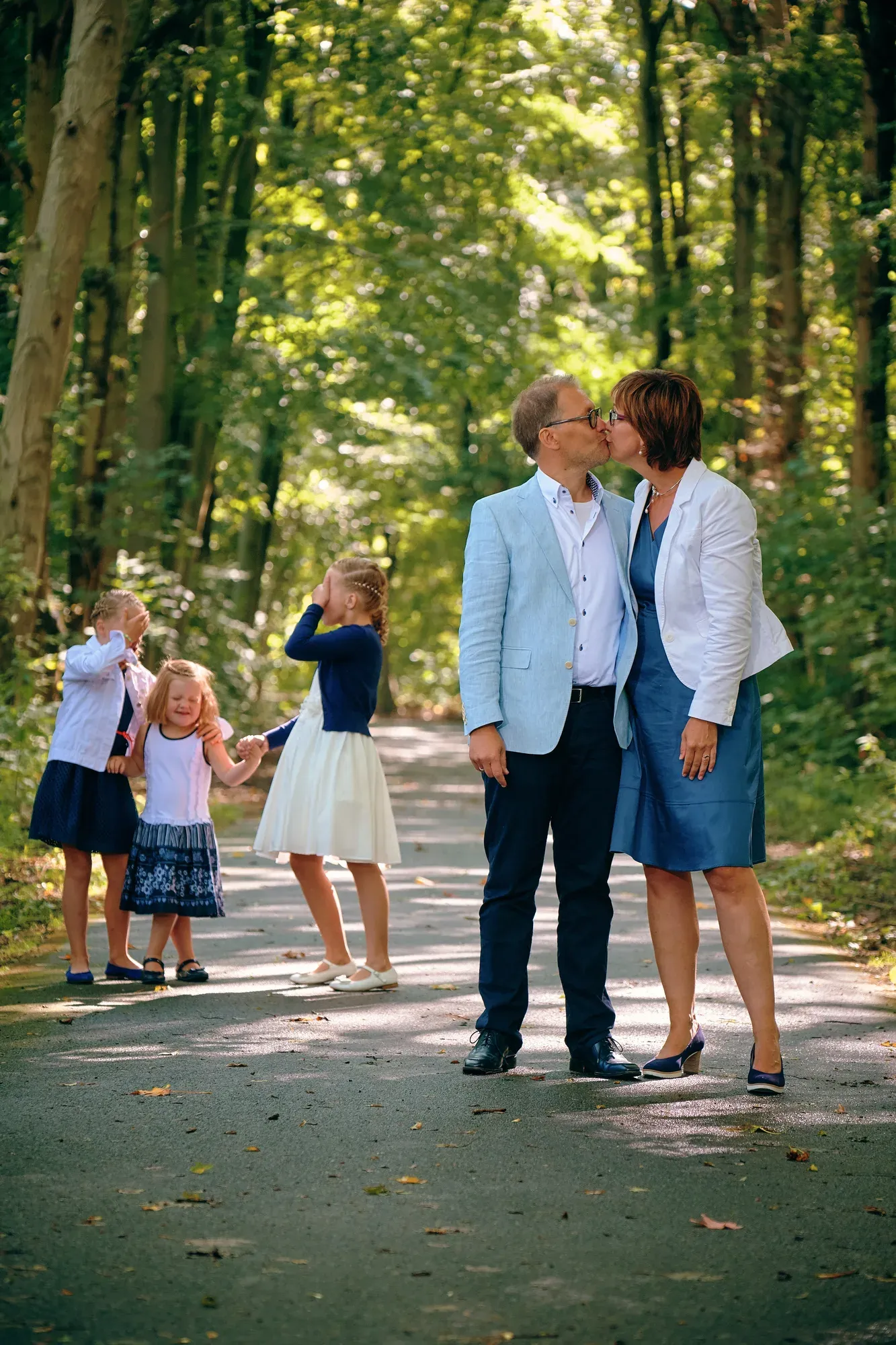 Ouders kussen elkaar op een bospad onder hoge bomen terwijl hun drie dochters op de achtergrond spelen