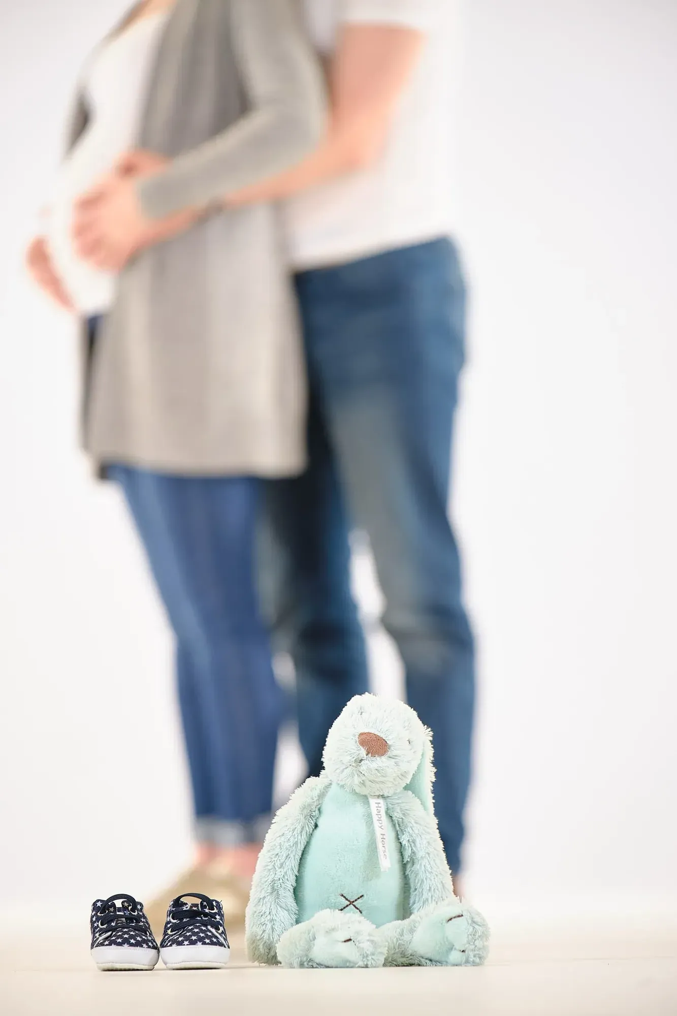 Zwanger stel staat samen in de witte studio, met een knuffel aan hun voeten
