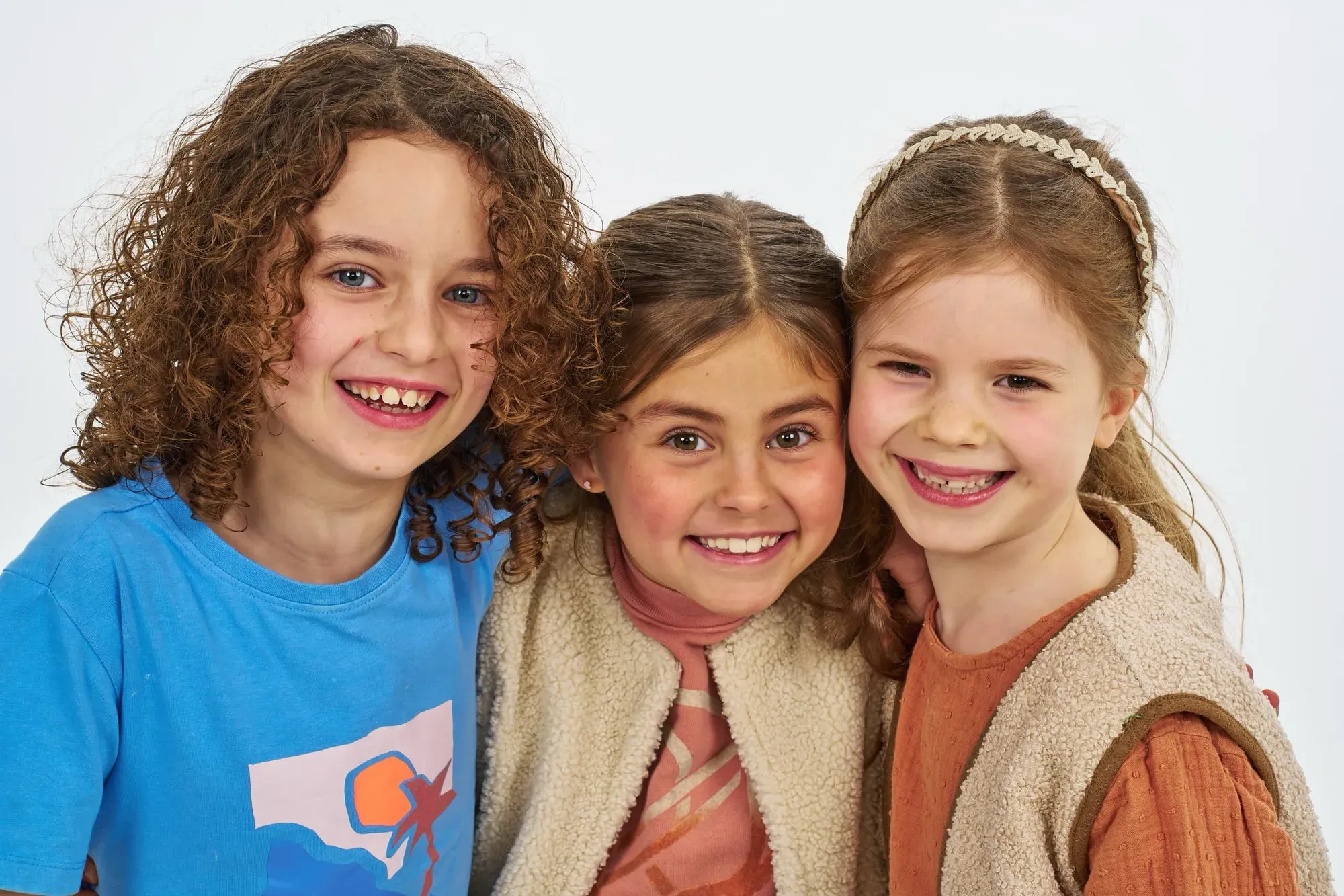 Drie zusjes met krullen staan lachend dicht tegen elkaar aan