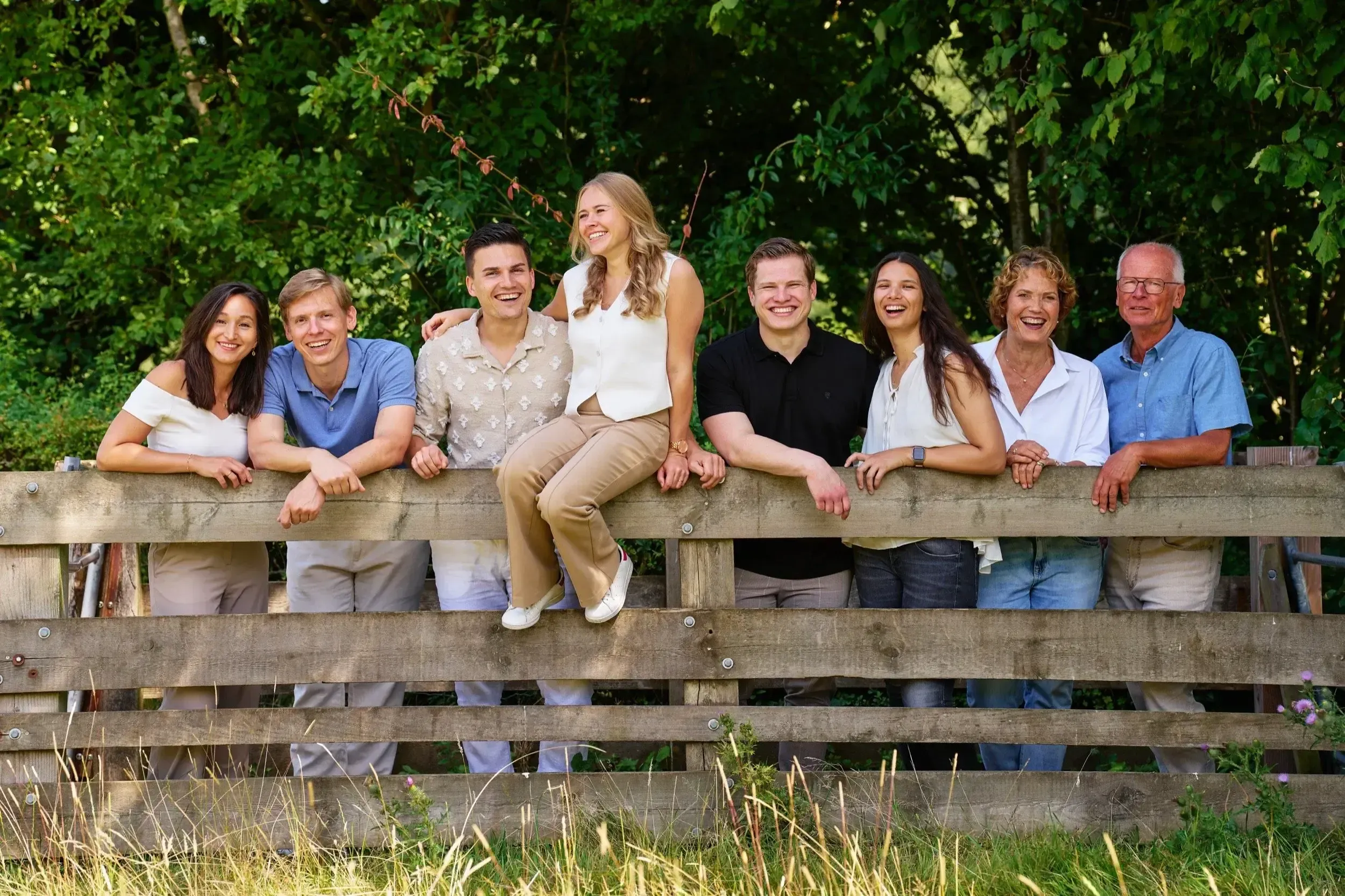 Groep van negen mensen die lachen en samen op een houten hek zitten, met groene bomen op de achtergrond.