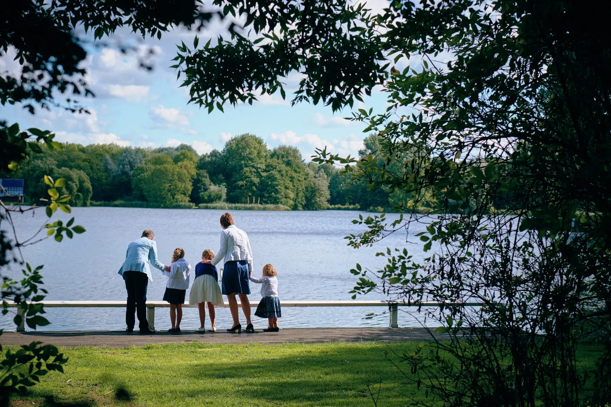 Gezin staat met de rug naar de camera aan het water, omlijst door de bladeren van de bomen