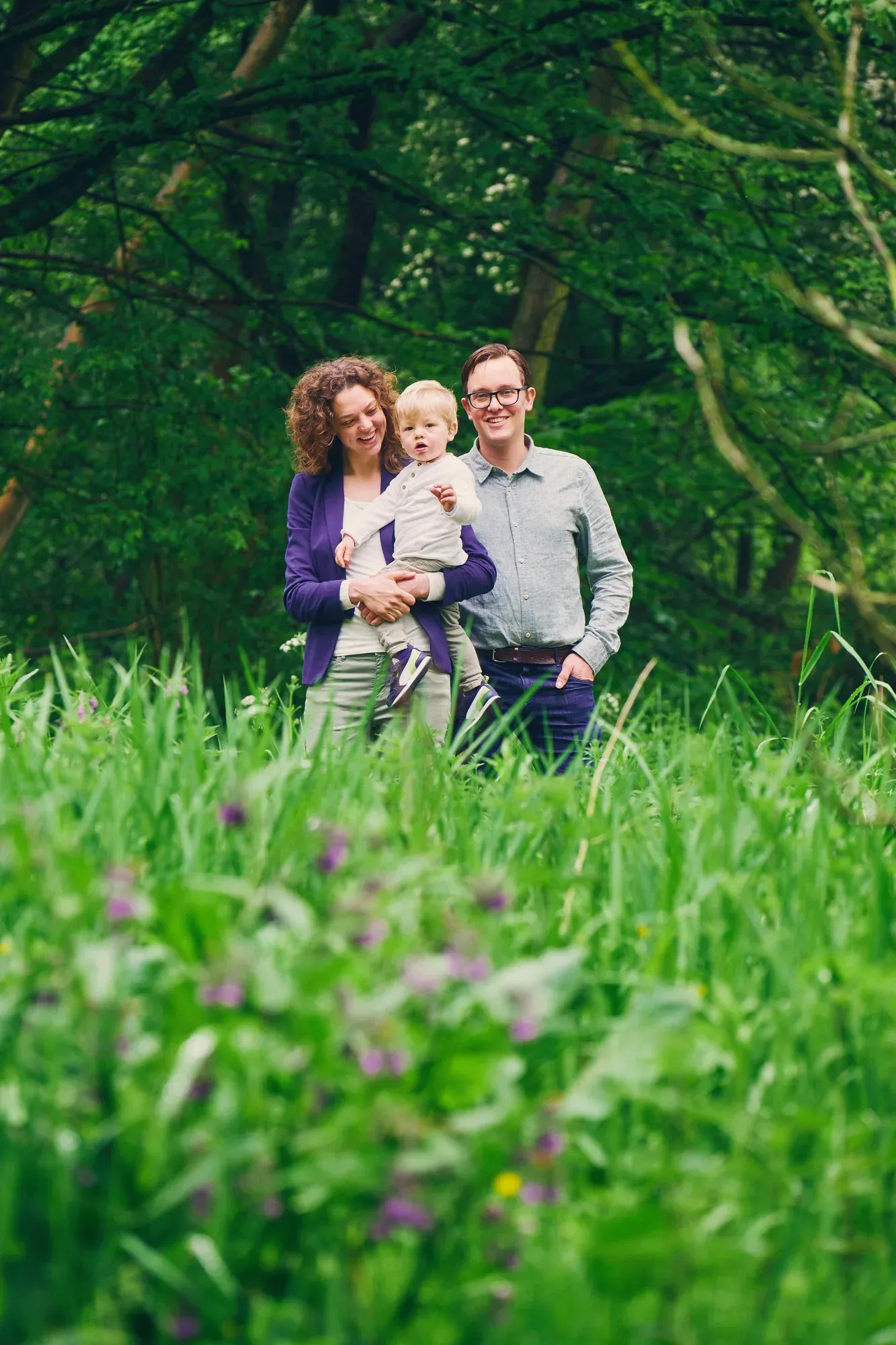 Ouders met peuter op de arm staan in hoog gras en wilde bloemen aan de rand van het bos