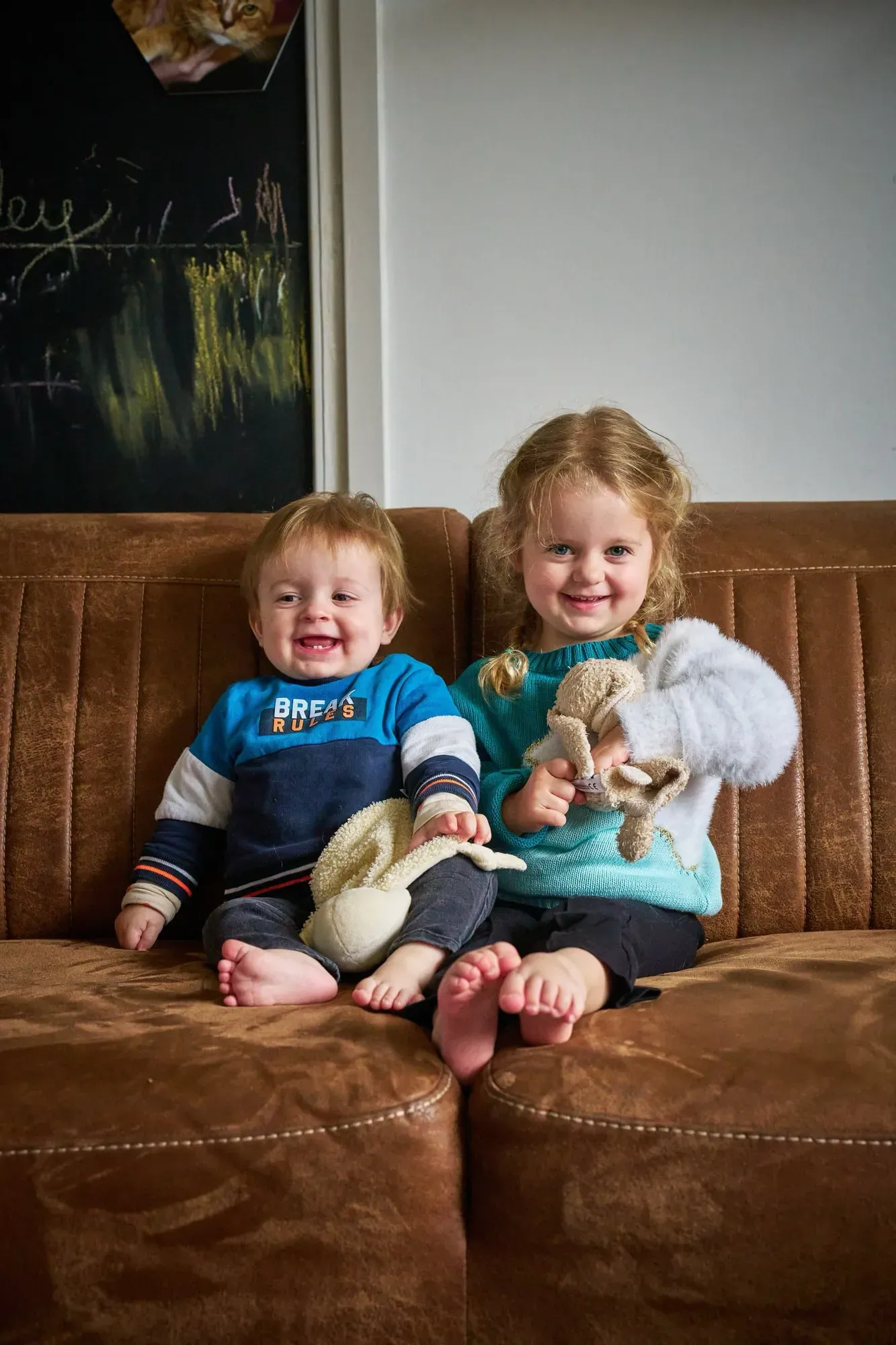 Twee kleine kinderen zitten lachend op een bruine bank met een knuffel