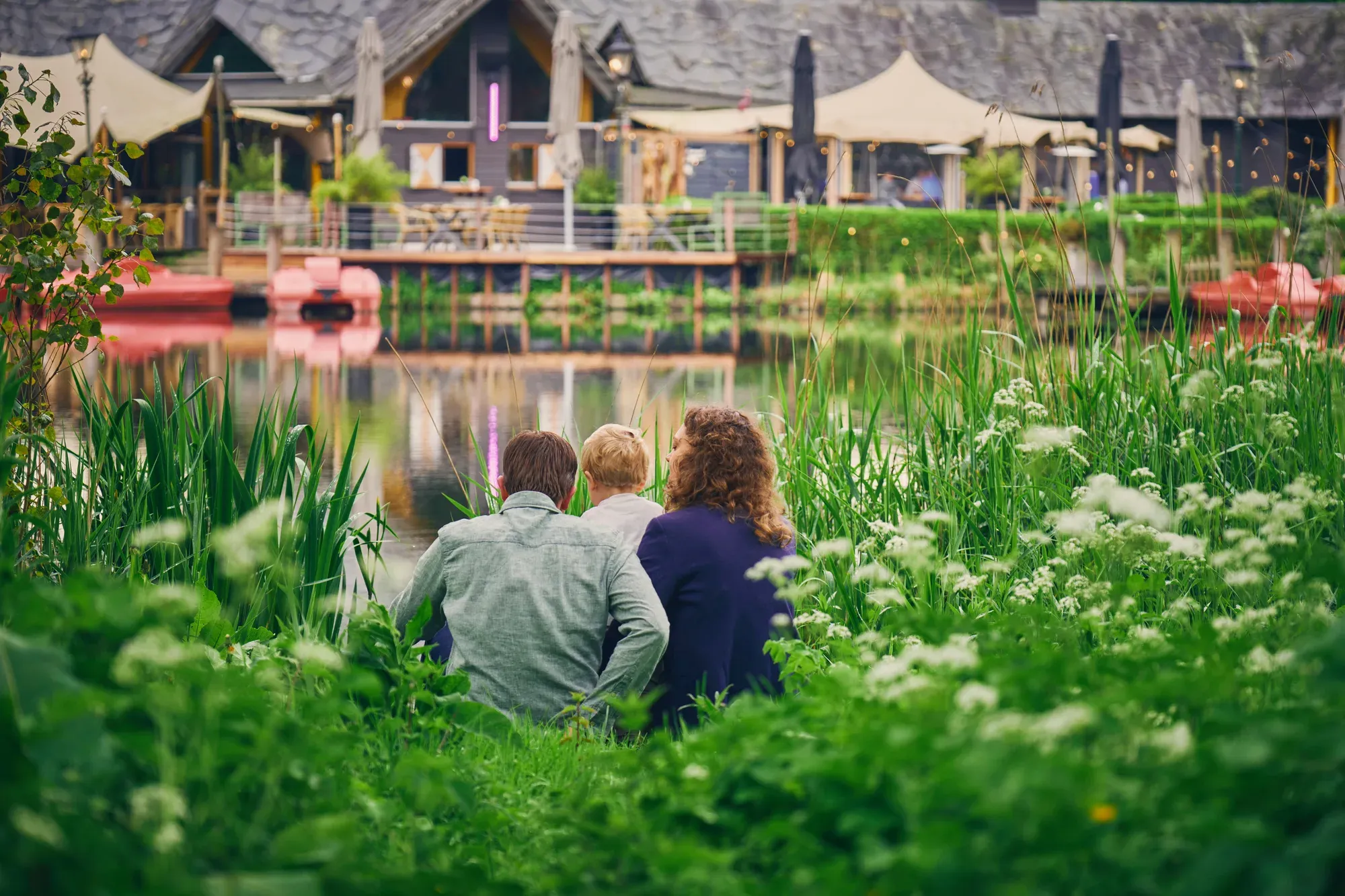 Ouders en peuter zitten met de rug naar de camera tussen het groen en kijken over het water naar Knus