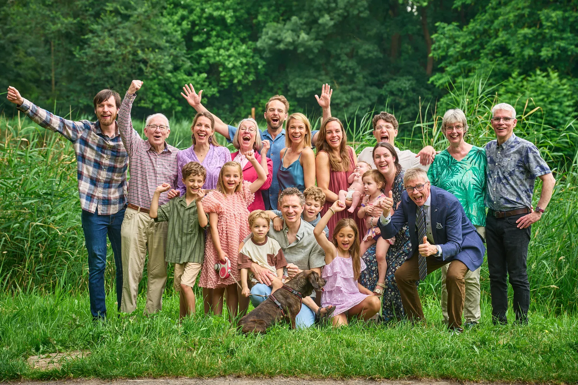 Juichende groepsfoto van een grote familie met drie generaties en een hond op een grasveld aan de bosrand